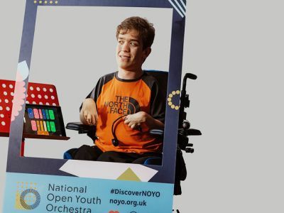 A boy wearing an orange North Face t-shirt and sat in a wheelchair sits in between a customised National Open Youth Orchestra Instagram frame